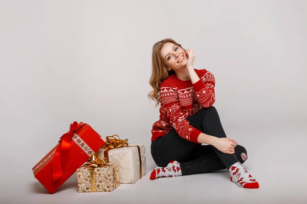 Stilvolle schöne junge Frau in einem modischen roten Strickpullover sitzt neben Geschenken auf grauem Hintergrund — Stockfoto