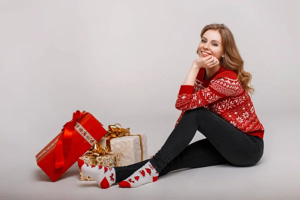 Mujer joven feliz en suéter rojo de moda con calcetines sentados cerca de regalos en el estudio —  Fotos de Stock