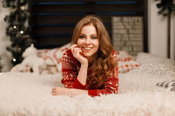 Bastante joven mujer feliz en un suéter de moda roja se encuentra en la cama y disfruta del momento —  Fotos de Stock