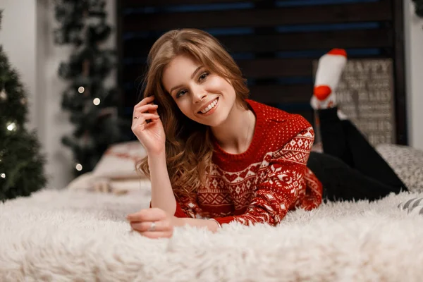 Feliz mujer hermosa a la moda en un suéter rojo con un adorno que descansa en una cama —  Fotos de Stock