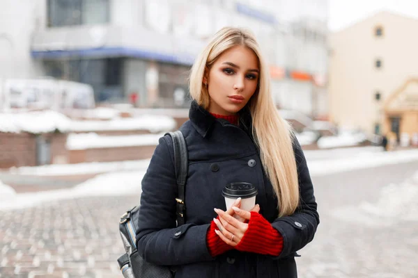 Menina da moda bonita com café em um casaco de inverno na moda com um saco andando na cidade — Fotografia de Stock