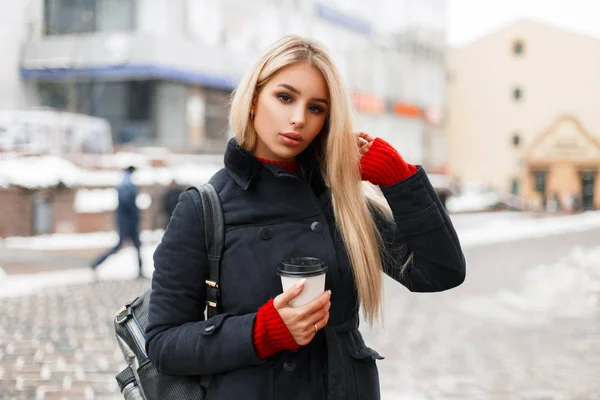 Jolie jeune femme blonde mannequin avec café dans un manteau de mode avec un sac dans la rue dans la ville — Photo