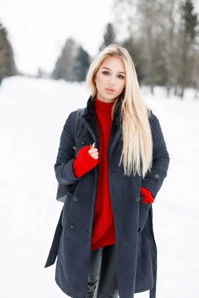 Mulher modelo bonita elegante em um casaco elegante de moda com uma camisola de malha vermelha na moda andando no parque em um dia de inverno — Fotografia de Stock