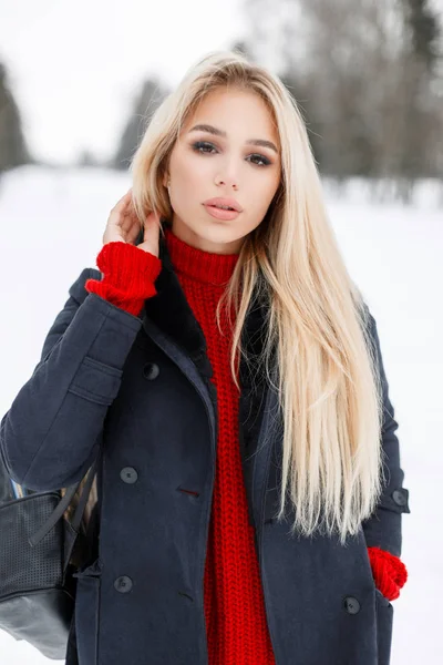 Hermosa chica con estilo joven en un abrigo de invierno de moda con un suéter rojo de punto caminando al aire libre en un día nevado — Foto de Stock