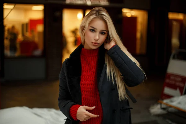 赤いセーターのエレガントなコートのライトと店の窓近くポーズ ファッションでかなり若い女性 — ストック写真