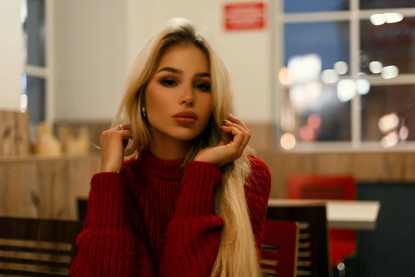 Piękna młoda kobieta modne w czerwonym swetrze siedzi przy stoliku w kawiarni w nocy — Zdjęcie stockowe