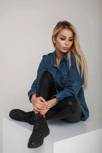 Americana bela jovem loira em roupas jeans com calças de couro em tênis preto elegante senta-se em estúdio — Fotografia de Stock