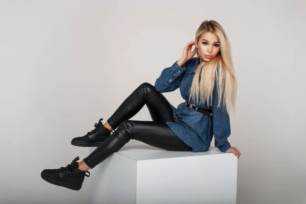 Красива молода американська модель жінка в джинсовому одязі з стильний чорний кросівки сидить у студії — стокове фото