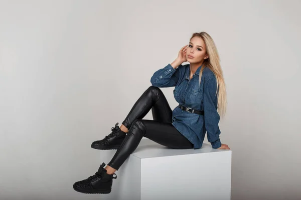 Молодая модная блондинка в джинсовой рубашке с черными стильными кроссовками позирует в студии — стоковое фото