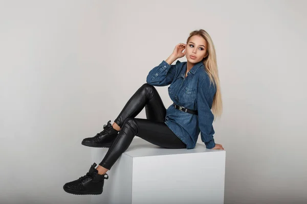 Молодая американская модель женщина в джинсовой рубашке и черных брюках с черными модными кроссовками сидит в студии — стоковое фото