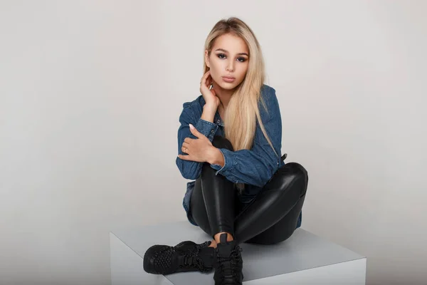 Güzel sexy genç kız moda kot giysileri siyah spor ayakkabı ile bir saç modeli ile stüdyoda oturur — Stok fotoğraf