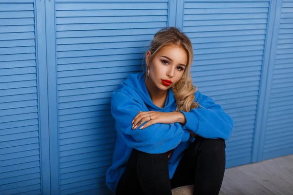 Élégante jeune femme en sweat à capuche tendance assise près d'un mur en bois bleu à l'intérieur — Photo