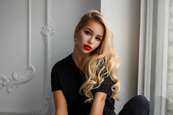 Bella donna alla moda modello giovane con un'acconciatura in una t-shirt nera seduta vicino a una parete vintage bianca — Foto Stock