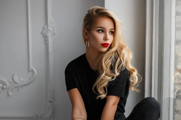 Hermosa mujer joven con estilo en una camiseta negra de moda sentado y mirando por la ventana —  Fotos de Stock