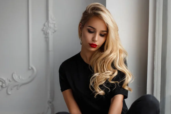 Елегантна модна молода жінка з зачіскою в стильній чорній футболці, що сидить біля білої стіни — стокове фото