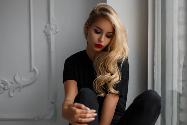 Ganska ung blond kvinna med make-up i svart mode T-shirt sitter nära fönstret — Stockfoto