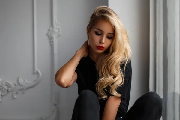 Joven mujer hermosa de moda con un peinado en una elegante camiseta negra sentada cerca de una pared blanca — Foto de Stock