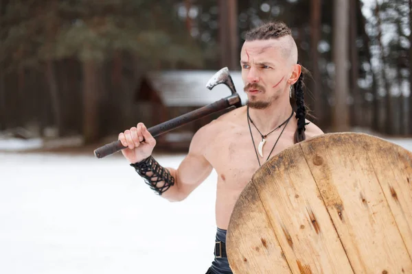 Viking man with a scar with an ax and a shield in the woods on a winter day