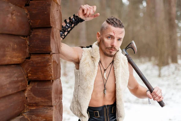 Viking com uma cicatriz com um machado em um colete de lã perto de uma parede de madeira na floresta em um dia de inverno — Fotografia de Stock