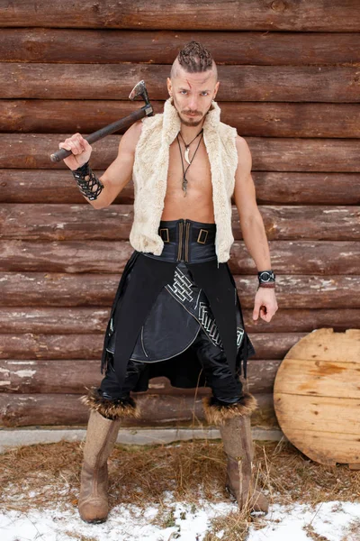Guerreiro Viking com um machado em um colete feito de pele em botas perto de uma parede de madeira — Fotografia de Stock