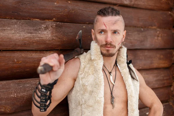Viking com uma cicatriz com um penteado segurar um machado em um colete de pele fica perto de uma parede de madeira — Fotografia de Stock