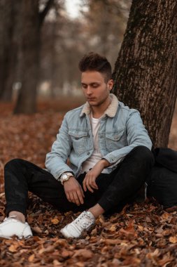 Beyaz spor ayakkabılarıyla şık giyinmiş genç yakışıklı hippi parktaki bir ağacın yanında yaprakların üzerinde dinleniyor. Çekici bir adam sonbahar manzarasından hoşlanıyor. Moda erkek kıyafetleri.