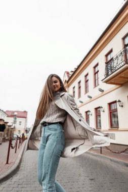 Moda trençkotlu, şık örülmüş süveter giyen, şehirde klasik bir binanın yanında dönen neşeli genç bir kadın modeli. Çekici, mutlu, modacı kız sokaklarda dolaşıyor..