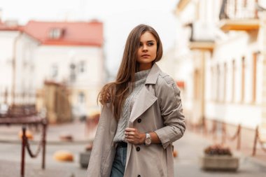 Şık bej renkli trençkotlu şık bir kadın modeli. Uzun kahverengi saçlı, şık örgü süveterli parlak bir bahar gününde şehirde dolaşıyor. Modern, sevimli bir kız. Gençlik modası.