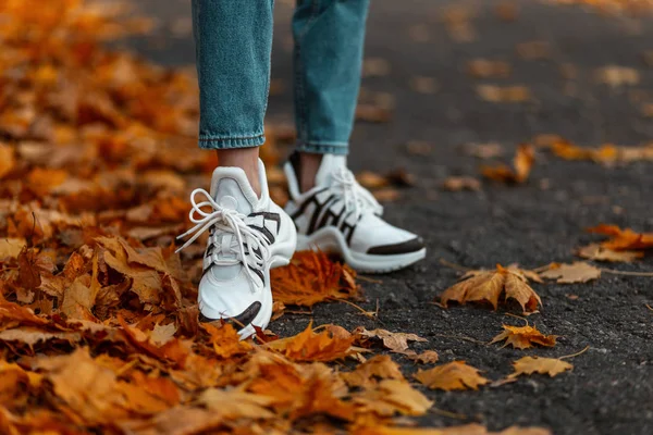 Módní bílé kožené tenisky. Detailní záběr ženských nohou ve stylově modrých džínách v módních botách. Moderní dívka procházky v parku na podzim den. — Stock fotografie