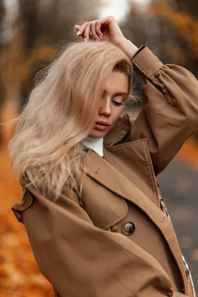 Portrait of a sensual young woman with blond hair in an elegant beige cloak in an autumn park on a background of trees with orange foliage. Attractive cute girl model posing and looking down outdoors. — Stockfoto