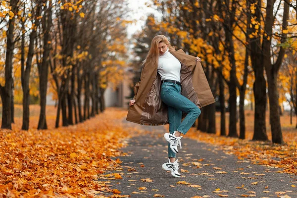 Docela elegantní mladá blondýnka ve stylovém kabátě v modrých džínách v módních teniskách pózující v uličce v parku na pozadí jasně oranžových stromů. Roztomilá holka pózující v lese. Podzimní móda — Stock fotografie