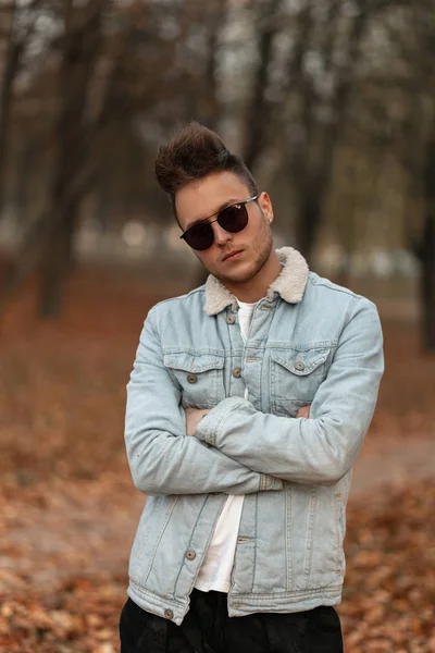 Modelo fresco joven americano en gafas de sol de moda en una chaqueta de mezclilla elegante con un peinado de moda en el parque. Chico guapo hipster posando en el bosque sobre un fondo de follaje otoñal . — Foto de Stock