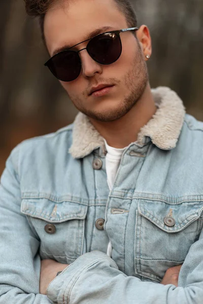 Retrato de un hombre hipster joven y elegante con gafas de sol en una chaqueta vaquera azul vintage con un peinado de moda al aire libre en un parque. Muchacho atractivo modelo de moda para un paseo en el bosque de otoño . — Foto de Stock