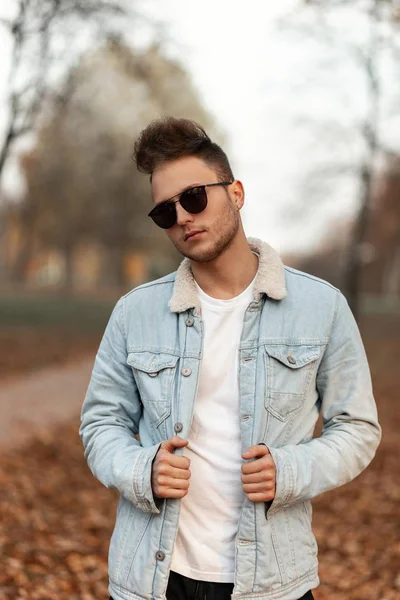 Homem moderno e bonito hipster jovem com penteado elegante em t-shirt em jaqueta de ganga vintage em óculos de sol pretos posa no parque em um dia de outono. Atraente na moda cara está andando na floresta . — Fotografia de Stock