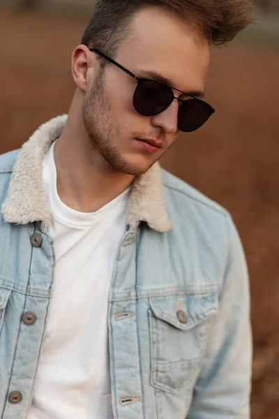 Portrait of a young handsome man with a beard with a fashionable hairstyle in stylish sunglasses in a white t-shirt in a denim jacket in the park. Attractive hipster guy is relaxes outdoors in nature. — Stockfoto