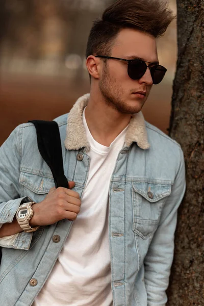 Un joven urbano con un peinado de moda en una elegante chaqueta de mezclilla azul con gafas de sol negras está descansando cerca de un árbol en un parque. Guapo hipster chico en ropa de moda está disfrutando del descanso en el bosque . — Foto de Stock