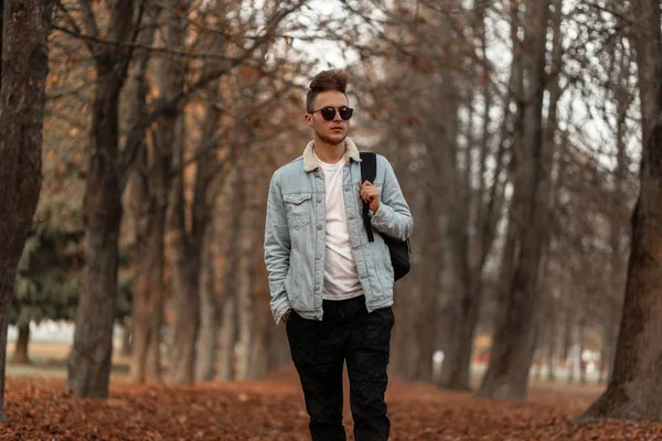 Trendy fiatal hipster férfi egy elegáns frizura egy vintage kék farmer kabát nadrágban napszemüvegben egy hátizsák utazik az őszi parkban egy októberi napon. Jóképű srác sétál az erdőben.. — Stock Fotó