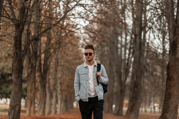 Europese trendy knappe jongeman in stijlvolle jeans kleding in vintage zonnebril met een modieus kapsel met rugzak wandelingen op het herfstbos. Aantrekkelijke kerel geniet van een wandeling in het park. — Stockfoto