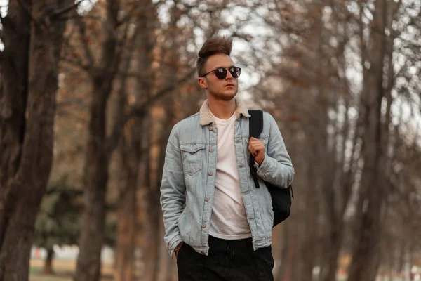 Américain élégant jeune homme hipster avec une coiffure à la mode dans des vêtements à la mode avec un sac à dos noir dans des lunettes de soleil à la mode se déplace à travers un parc d'automne sur un fond d'arbres. . — Photo