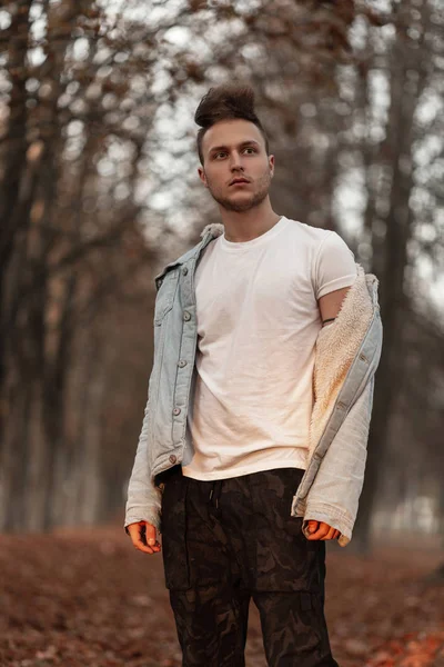 Joven guapo joven de moda en una camiseta blanca de moda en una elegante chaqueta de mezclilla azul con un peinado posando en el parque de otoño. Atractivo tipo europeo al aire libre. Moda masculina de moda juvenil . — Foto de Stock