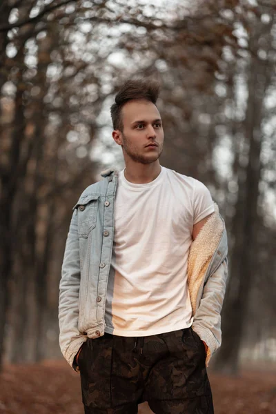 Knappe moderne jongeman in een vintage wit T-shirt in een blauw denim jasje met een modieus kapsel dat buiten poseert in een park. Trendy vent rustend in de natuur op een herfstdag. Stijlvolle herenkleding. — Stockfoto