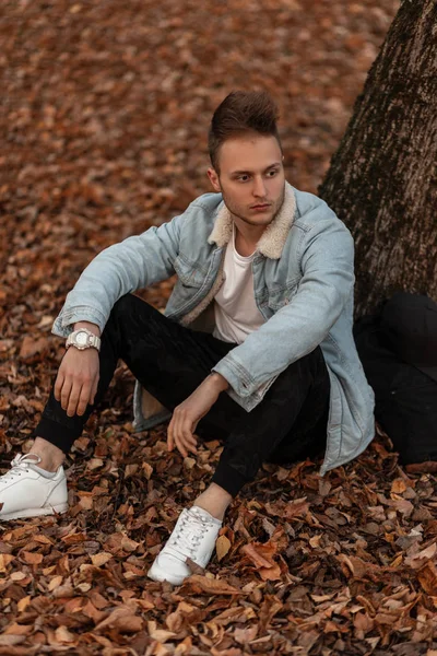 Niza joven americano en pantalones militares con estilo en una chaqueta de mezclilla de moda vintage en zapatillas blancas de moda se sienta en el follaje seco dorado en un parque. Modelo chico de moda se relaja en el bosque . — Foto de Stock