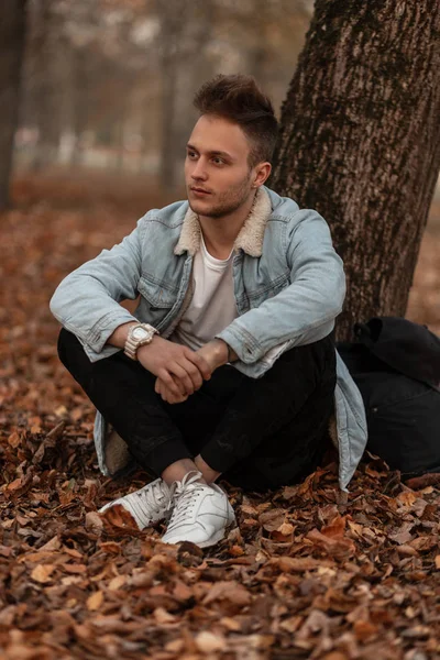 Joven hombre agradable de moda con un peinado elegante con barba en ropa de moda está descansando al aire libre en un parque sentado en el follaje naranja cerca de un árbol. Guapo chico modelo relaja en el bosque . — Foto de Stock