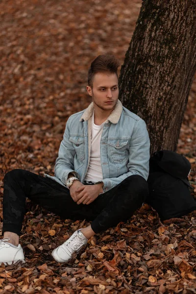 Modelo de un joven atractivo en una elegante chaqueta de mezclilla en zapatos de gimnasio en pantalones militares con una mochila con una barba con un corte de pelo cerca de un árbol en el parque. Cool chico está sentado en el follaje de otoño . — Foto de Stock