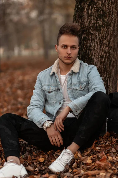 Un joven europeo con una elegante chaqueta de mezclilla en pantalones militares de moda en zapatillas blancas con una mochila está descansando cerca de un árbol en el bosque de otoño. Buen tipo sentado en el follaje dorado en un parque . —  Fotos de Stock