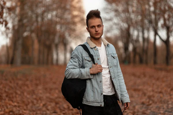 Fiatal jóképű férfi, divatos frizurával, elegáns pólóban, vintage farmerdzsekiben, hátizsákkal áll a parkban, arany lombozat hátterében. Vonzó fickó az őszi erdőben.. — Stock Fotó