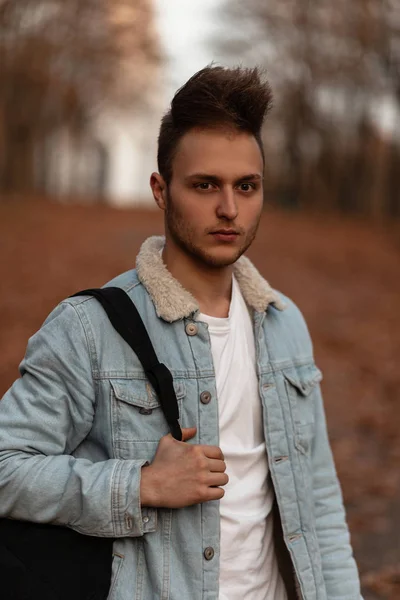 Retrato de un joven guapo con un peinado de moda en una chaqueta de mezclilla de moda en una camiseta con una mochila en el bosque de otoño. Un tipo atractivo camina por el parque en un cálido día de octubre. Estilo . —  Fotos de Stock
