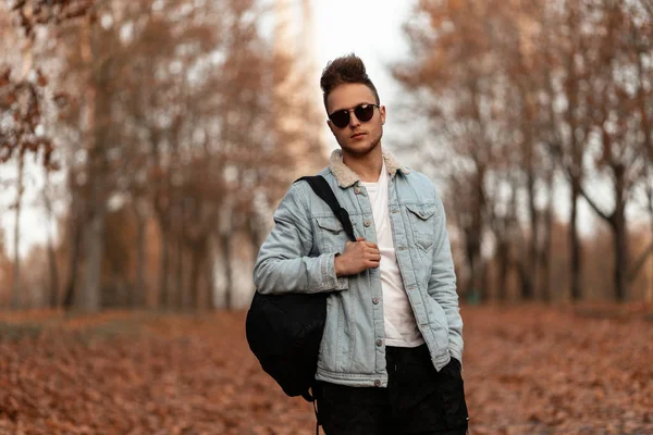 Hombre hipster joven guapo fresco con mochila con peinado elegante en camiseta en chaqueta de mezclilla vintage en gafas de sol negras posa en el parque en un día de otoño. Atractivo chico de moda en el bosque . — Foto de Stock