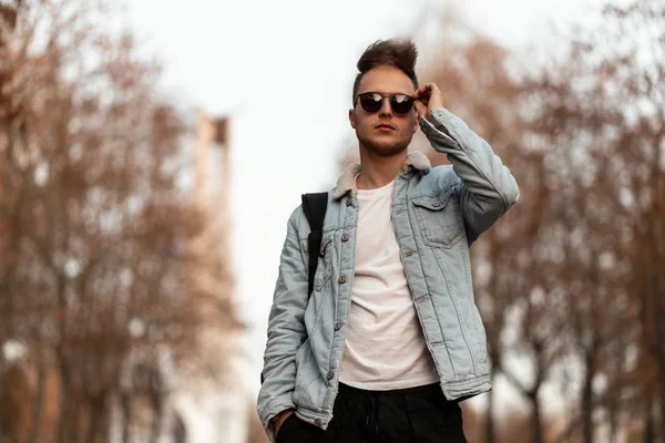 Stylish young man with a beard with a fashionable hairstyle in trendy sunglasses in a blue denim jacket is relaxing outdoors in the park on an October day. Handsome hipster guy. Youth autumn fashion. — Stock Photo, Image