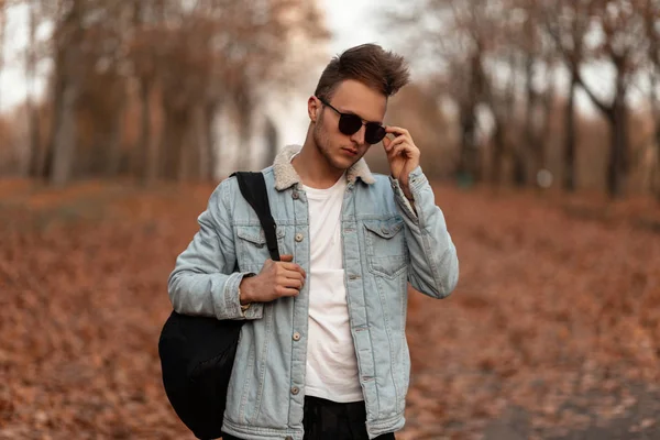 Jeune homme moderne à la mode avec une coiffure élégante en lunettes de soleil noires dans une veste en denim avec un sac à dos posant dans la forêt d'automne. Attrayant hipster guy en vêtements à la mode à l'extérieur dans une forêt . — Photo